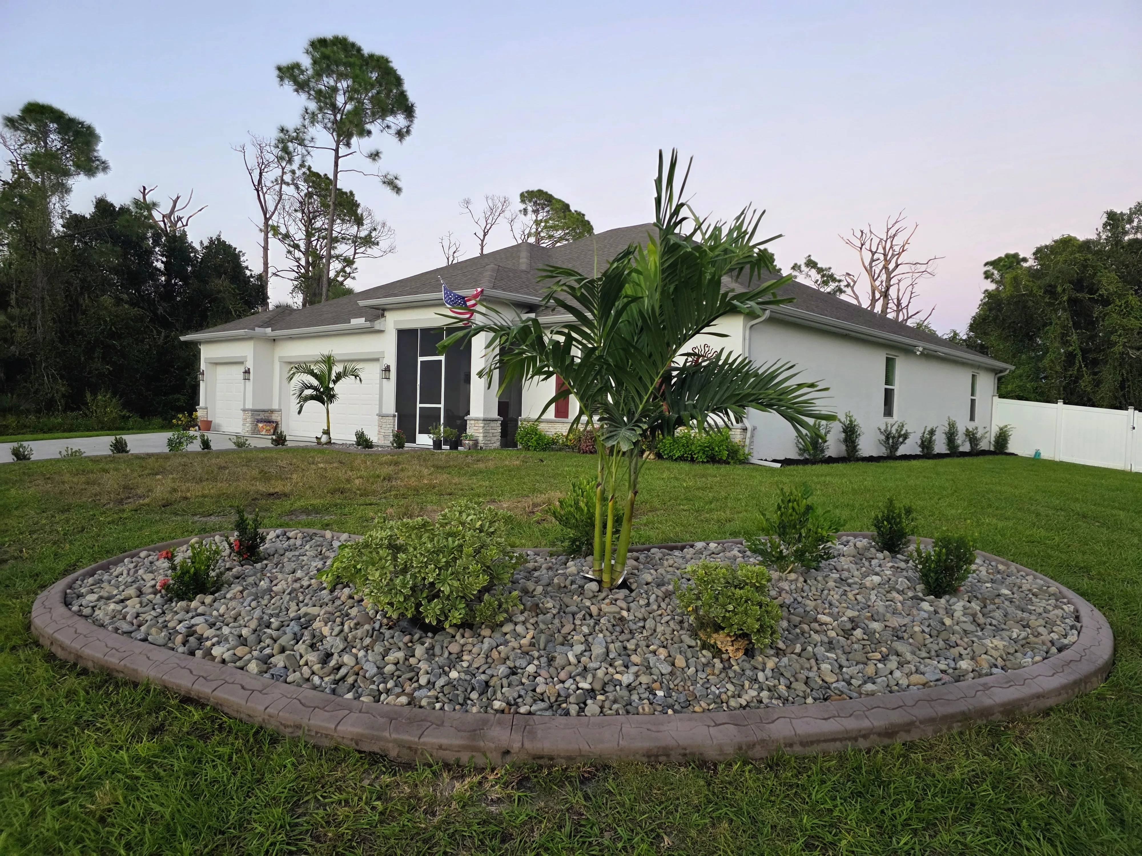 Oval island bed with brown stamped concrete curbing, river rock, Christmas palm and shrubs in a Florida front lawn