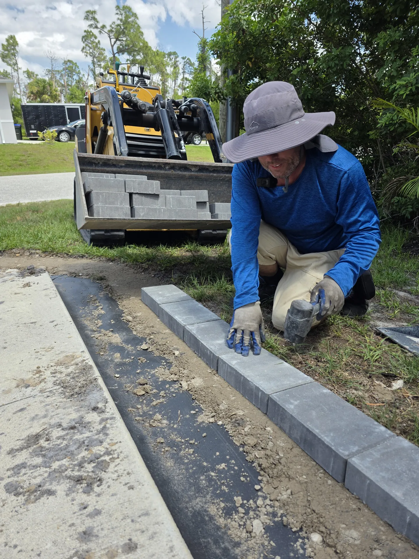 Worker hand-setting grey paver border edging along a driveway with a loaded skid steer behind