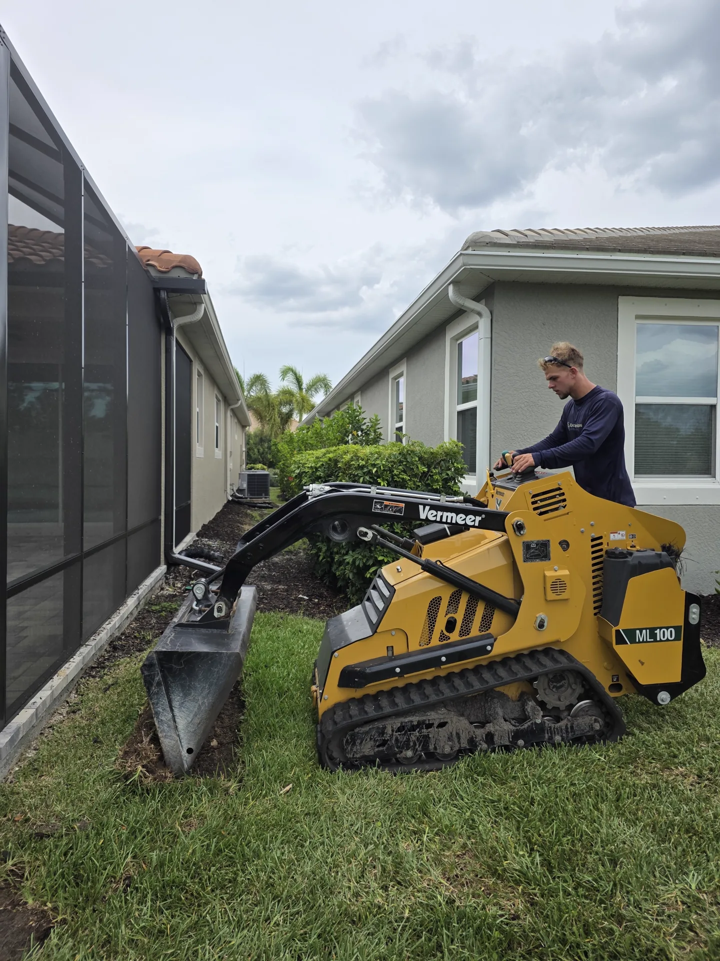 Epic Horizons operator clearing a side-yard bed with a compact tracked mini skid steer between two Southwest Florida homes