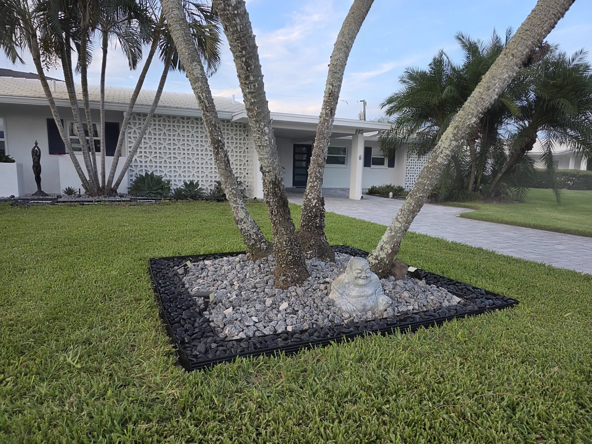 Square rock bed of gray granite with a black pebble border and steel edging ringing a cluster of palm trunks in a manicured lawn