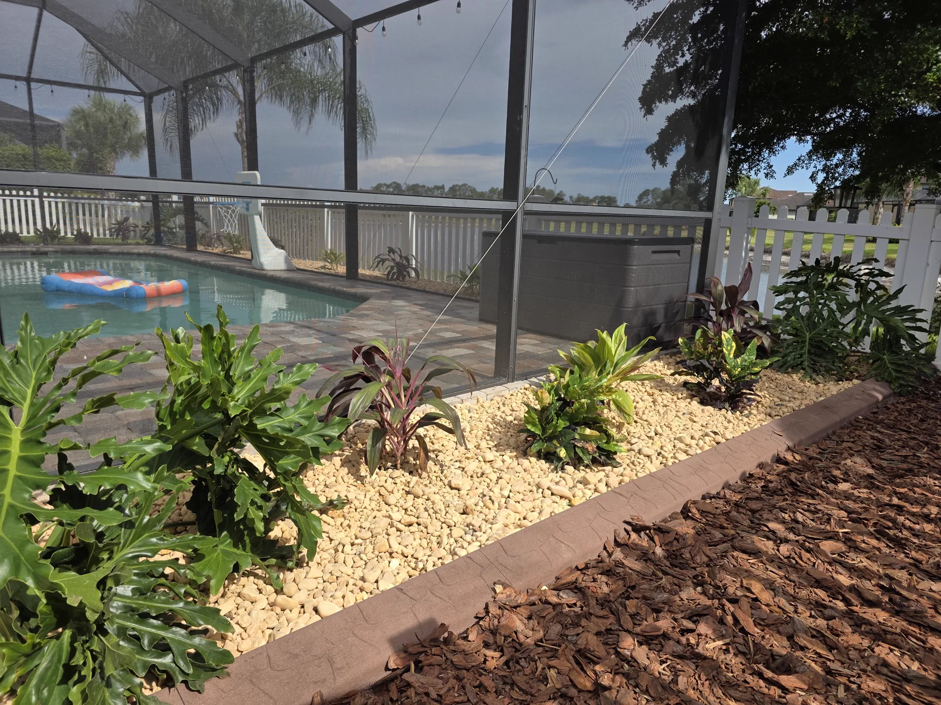 Brown stamped concrete curbing edging a white river rock planting bed with tropical plants beside a screened paver pool deck